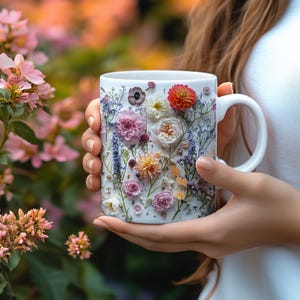 Op de afbeelding: Een witte keramische mok met een bloemenmotief. De mok is versierd met geperste bloemen in tinten roze, paars, wit en geel. De mok wordt vastgehouden door een hand.