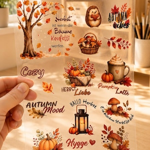 Könnte beinhalten: Ein transparenter Aufkleberbogen mit verschiedenen herbstlichen Illustrationen wie einem Baum mit orangefarbenen Blättern, einem Igel, einem Korb mit Pilzen, einem Kürbis, einer Laterne und einer Tasse, ergänzt durch Texte wie Herbstliebe.