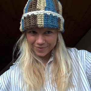 Könnte beinhalten: Eine handgefertigte Häkelmütze in einem gestreiften Muster aus Braun, Blau und Beige. Der Hut hat einen weißen, gewellten Rand. Die Person, die den Hut trägt, trägt auch ein weiß-blau gestreiftes Hemd.