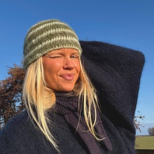 Könnte beinhalten: Eine Person trägt eine grün-weiß gestreifte Strickmütze, einen dunkelblauen Schal und einen dunkelblauen Mantel. Der Hintergrund ist ein klarer blauer Himmel.