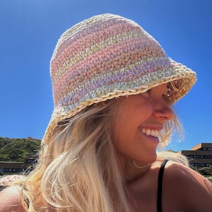 Könnte beinhalten: Eine Frau trägt einen gehäkelten Bucket-Hut mit rosa, grünen und weißen Streifen. Der Hut hat eine breite Krempe und ist aus einem weichen, strukturierten Garn gefertigt.