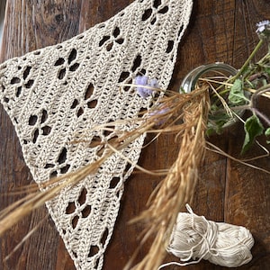 Könnte beinhalten: Ein dreieckiges, cremeweißes Häkelbandana mit einem sich wiederholenden Durchbruchmuster aus kleinen vierblättrigen Blumenmotiven. Das Stück ist aus hellem Garn gefertigt und weist eine zarte, luftige Textur in seinem geometrischen Design auf.
