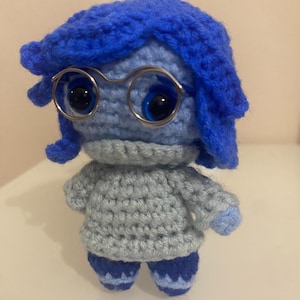 Può includere: Bambola all'uncinetto fatta a mano con un personaggio con capelli blu, occhiali e un maglione grigio. La bambola ha grandi occhi blu e indossa un completo blu. La bambola è alta circa 13 cm.