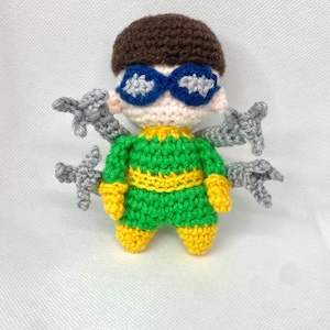 Può includere: Bambola amigurumi fatta a mano del Dottor Octopus. La bambola presenta una testa marrone, occhiali blu e bianchi e un completo verde e giallo. Quattro braccia meccaniche grigie si estendono dalla schiena della bambola. La bambola è fatta di filato.