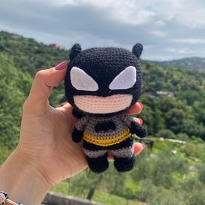 Può includere: Un giocattolo Batman all'uncinetto con un mantello nero, un vestito grigio e una cintura gialla. Il giocattolo è tenuto in una mano su uno sfondo di alberi verdi e un cielo nuvoloso.