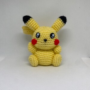 Può includere: Un peluche Pikachu di colore giallo lavorato a uncinetto con orecchie nere e guance rosse. Il giocattolo ha occhi neri e un piccolo naso nero.