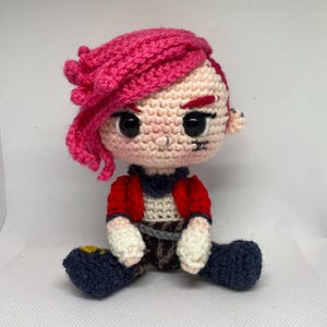 Puede incluir: Muñeca de ganchillo hecha a mano con pelo rosa, ojos negros y un pequeño piercing en la nariz. La muñeca lleva una chaqueta roja, una camiseta azul y blanca y pantalones azules. La muñeca está sentada con las piernas dobladas y los brazos a los lados.
