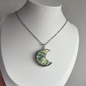 May include: A crescent moon-shaped labradorite pendant wrapped in silver-toned wire, suspended from a thin silver-toned metal chain. The stone displays iridescent green and blue flashes across its polished surface, secured by a wire bail.