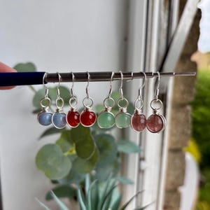 Puede incluir: Una colección de pendientes de piedras preciosas en varios colores, incluyendo azul, rojo y verde. Cada pendiente presenta una pequeña piedra redonda envuelta en alambre de plata y suspendida de un gancho de plata. Los pendientes se exhiben en una varilla metálica.