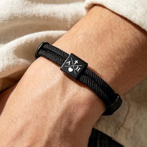 May include: A black double-strand cord bracelet featuring a square-shaped central charm. The charm is engraved with a cross symbol, a heart icon, and the letters A and H arranged in a cross pattern on its matte black surface.