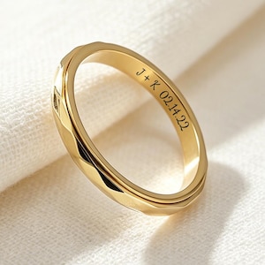 May include: A gold ring with a faceted design around the band. The inside of the ring is engraved with the initials "J+K" and the date "02.14.22". The ring is set against a white fabric background.