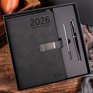 Może przedstawiać: Czarny, skórzany notatnik z harmonogramem na rok 2026 ze srebrnym zapięciem, prezentowany w pudełku prezentowym z pasującym długopisem i rysikiem. Notatnik jest wytłoczony imieniem "Klein". Zestaw jest prezentowany na drewnianej powierzchni.