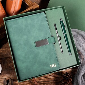 May include: A green leather journal with a metal clasp, presented in a matching gift box with a green pen and a refill. The journal is embossed with the initials "NG". The background includes a wooden box and a small plant.