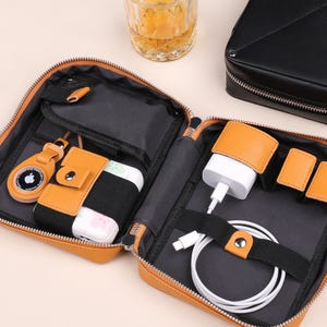 May include: Open, tan leather travel organiser with a zip, containing a charger, cables, and a small AirTag holder. The interior is black. A glass of amber liquid and a black leather case are in the background.