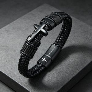 May include: A black braided bracelet featuring a central cross charm engraved with the name ANDERSON. The bracelet includes textured metal accents and a rectangular clasp engraved with the text NEVER ALONE and a small white cross symbol.