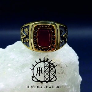 May include: A gold ring with a red stone set in a wreath design. The ring has black accents and a design on either side of the stone.