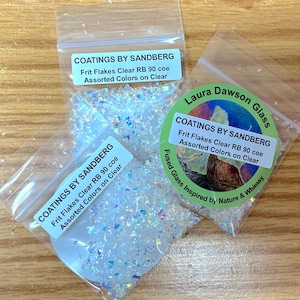 May include: Three clear plastic bags filled with iridescent glass frit flakes. Each bag has a label that reads "COATINGS BY SANDBERG" and "Frit Flakes Clear RB 90 coe Assorted Colors on Clear." One bag has a green circular label that says "Laura Dawson Glass."