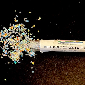 May include: A close-up of dichroic glass frit flakes in rainbow colors, scattered on a black surface. The flakes are spilling from a tube labeled "Dichroic Glass Frit Flakes Rainbow". The tube has a logo with a rainbow design.