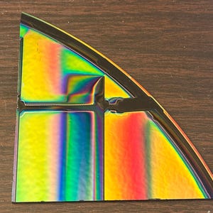 May include: A colorful, iridescent, triangular piece with a black border. The surface reflects a spectrum of colors, including yellow, green, blue, and red. The shape is a segment of a circle, with a cut-out section.