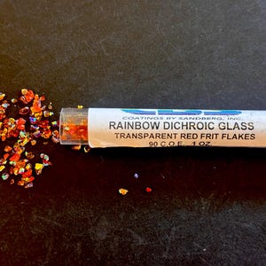 May include: A clear tube containing rainbow dichroic glass flakes, with some flakes scattered beside it. The label reads "RAINBOW DICHROIC GLASS TRANSPARENT RED FRIT FLAKES 90 C.O.E. 1 OZ." The flakes are a mix of red, orange, and yellow.