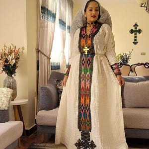 Puede incluir: Un vestido crema de manga larga con intrincados bordados en colores vibrantes. El vestido presenta un patrón detallado en la parte delantera y en las mangas. Un colgante de cruz dorada cuelga del escote. El vestido se usa con un velo a juego.