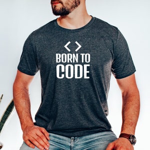 Op de afbeelding: Een donkergrijs T-shirt met witte tekst die "< > Born to Code" luidt. De persoon die het shirt draagt, zit met zijn handen op schoot.