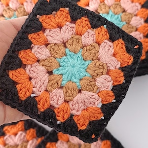 Könnte beinhalten: Nahaufnahme von mehreren bunten Häkelquadraten. Jedes Quadrat hat eine zentrale türkisfarbene Blumenform, umgeben von Blütenblättern in Rosa, Orange und Braun, alles eingerahmt von einem schwarzen Rand. Die Quadrate sind handgefertigt und wirken weich.