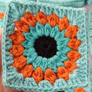 Peut inclure: Gros plan sur un carré crocheté avec un motif de tournesol. Le centre est noir, entouré de pétales turquoise et encadré de pétales orange. La bordure extérieure est turquoise. Plusieurs carrés similaires sont visibles en arrière-plan.