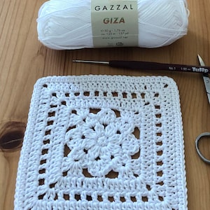 May include: A white crocheted square with a floral design in the center, a ball of white yarn, a crochet hook, and scissors. The yarn label reads "GAZZAL GIZA". The hook is a size No. 1, and the scissors are marked "Tulip".