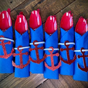 May include: Six nautical-themed place settings. Each setting includes a red spoon, blue napkin, a red glitter anchor, and a white rope napkin ring. The settings are arranged on a wooden surface.