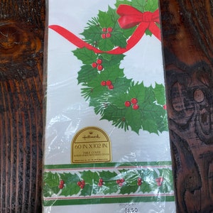 May include: A white plastic-wrapped table cover with a green holly wreath design and a red bow. The table cover is 60 inches by 102 inches and has a white center with a border design. The text "Hallmark" is printed on the cover. The price is $1.50.