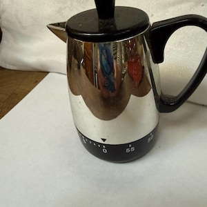 May include: A silver-colored metal coffee percolator with a black lid, black handle, and a black base featuring a built-in mechanical timer. The timer dial displays white numbers including 50 and 55, with a small downward-pointing triangle indicator.
