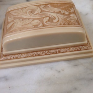 Pu&ograve; includere: Una scatola rettangolare color crema con coperchio, con intricate incisioni floreali in rilievo. Il coperchio e la base hanno un bordo decorativo. La scatola &egrave; posta su una superficie di marmo bianco, mostrando la lavorazione.
