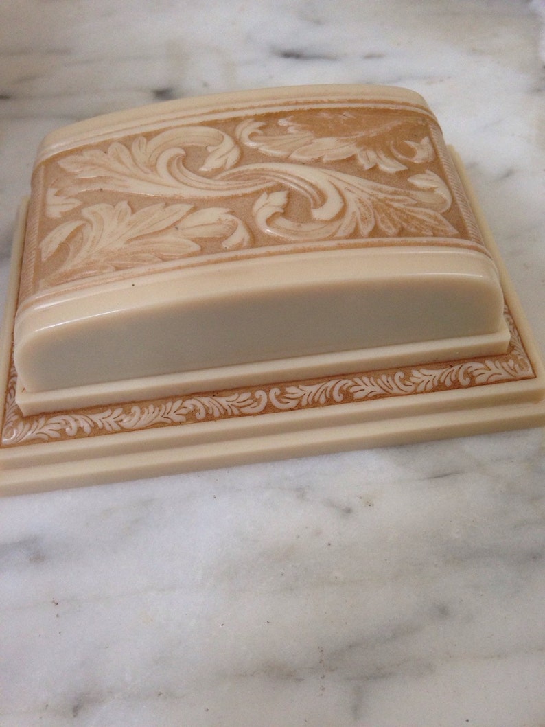 Pu&ograve; includere: Una scatola rettangolare color crema con coperchio, con intricate incisioni floreali in rilievo. Il coperchio e la base hanno un bordo decorativo. La scatola &egrave; posta su una superficie di marmo bianco, mostrando la lavorazione.