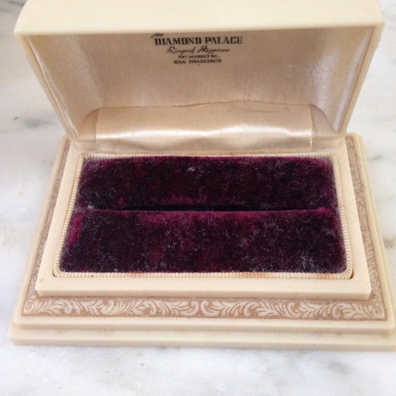 Pu&ograve; includere: Portagioie color avorio aperto con interno in velluto bordeaux scuro. La scatola &egrave; rettangolare con coperchio incernierato e presenta il testo "The Diamond Palace, Ring of Happiness, San Francisco" all'interno del coperchio.