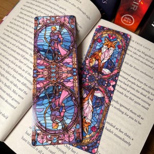 K&ouml;nnte beinhalten: Zwei Glas-Buchzeichen, eines mit einem Prinzen mit Bogen und Pfeil in einem blauen und rosafarbenen Glasrahmen, das andere mit einem Fuchs in einem rosafarbenen und blauen Glasrahmen.