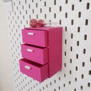 May include: A bright pink storage unit with three stacked drawers, each featuring a small white rectangular handle. The unit is mounted onto a white pegboard panel. Two round, multi-coloured striped lollipops rest on the top surface.