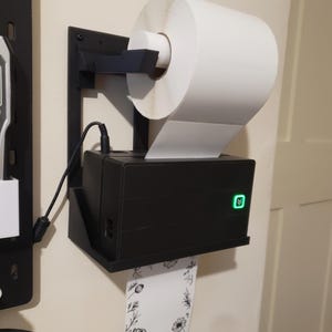 May include: A black label printer mounted on a wall, holding a roll of white labels. The printer has a green power indicator and is connected to a power source. A label with a floral design is dispensed.