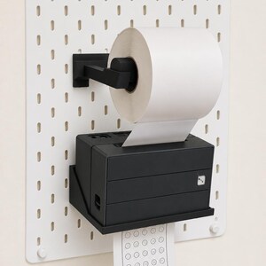 May include: A white pegboard wall mount holds a black paper roll dispenser and a black thermal printer. The printer outputs a white paper strip featuring a grid of numbered circles and the text StudyPpan with a small star and cartoon face icon.