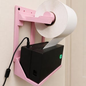 May include: A light pink wall-mounted plastic holder supporting a black thermal label printer and a large roll of white adhesive labels. The printer features a small green indicator light on the front and a black power cable connected to the top.