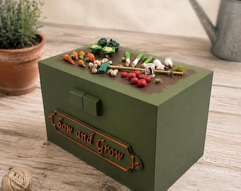 May include: A rectangular olive green wooden box with a hinged lid featuring miniature garden vegetables, including carrots, tomatoes, and greens. A copper-colored plaque on the front displays the embossed text "Sow and Grow" in a script font.