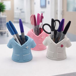 May include: Three pen holders in the shape of nurse uniforms in blue, pink, and white. Each has a cross and holds pens, markers, and scissors. The background is a blurred medical setting.
