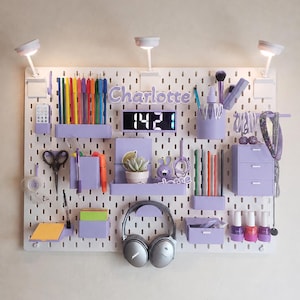 Peut inclure: Un panneau perforé blanc avec le prénom Charlotte en lettres violettes, une horloge numérique affichant 1421, trois petites lampes LED et divers bacs de rangement violets contenant des stylos, des ciseaux, un casque, du vernis et une plante.