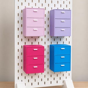 Puede incluir: Un panel perforado blanco con cuatro unidades de almacenamiento adjuntas, cada una con tres cajones. Los cajones son de color rosa claro, lavanda, rosa intenso y azul, todos con pequeñas manijas horizontales blancas.