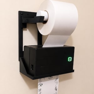 May include: A black wall-mounted thermal label printer with a roll of white paper loaded on top. The printer features a green illuminated power button with a double arrow icon. A strip of white paper with a black floral border design hangs down.