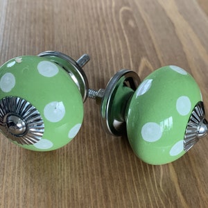 Op de afbeelding: Twee groene keramische kastknoppen met witte stippen en zilveren accenten. De knoppen zijn rond en hebben een verhoogd zilveren midden.