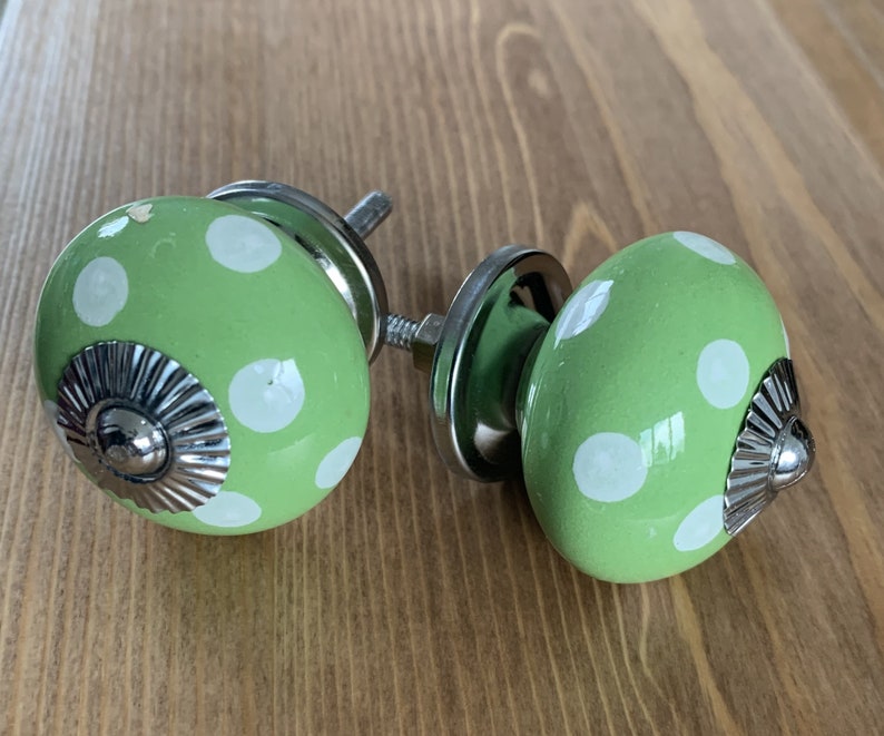 Op de afbeelding: Twee groene keramische kastknoppen met witte stippen en zilveren accenten. De knoppen zijn rond en hebben een verhoogd zilveren midden.