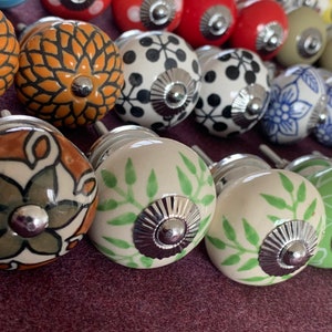 Op de afbeelding: Assortiment van keramische ladeknopjes met verschillende patronen en kleuren, waaronder groen, blauw, bruin, wit en zwart. De knoppen hebben een zilveren metalen accent in het midden.