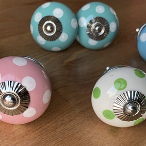 Op de afbeelding: Assortiment van zes keramische kastknoppen met een stippenpatroon. De knoppen zijn in verschillende kleuren, waaronder roze, blauw en groen. Elke knop heeft een zilveren metalen basis.