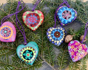 May include: Six handmade heart-shaped ornaments with colorful geometric patterns and purple cords. The ornaments are arranged on a dark gray surface with green pine branches.